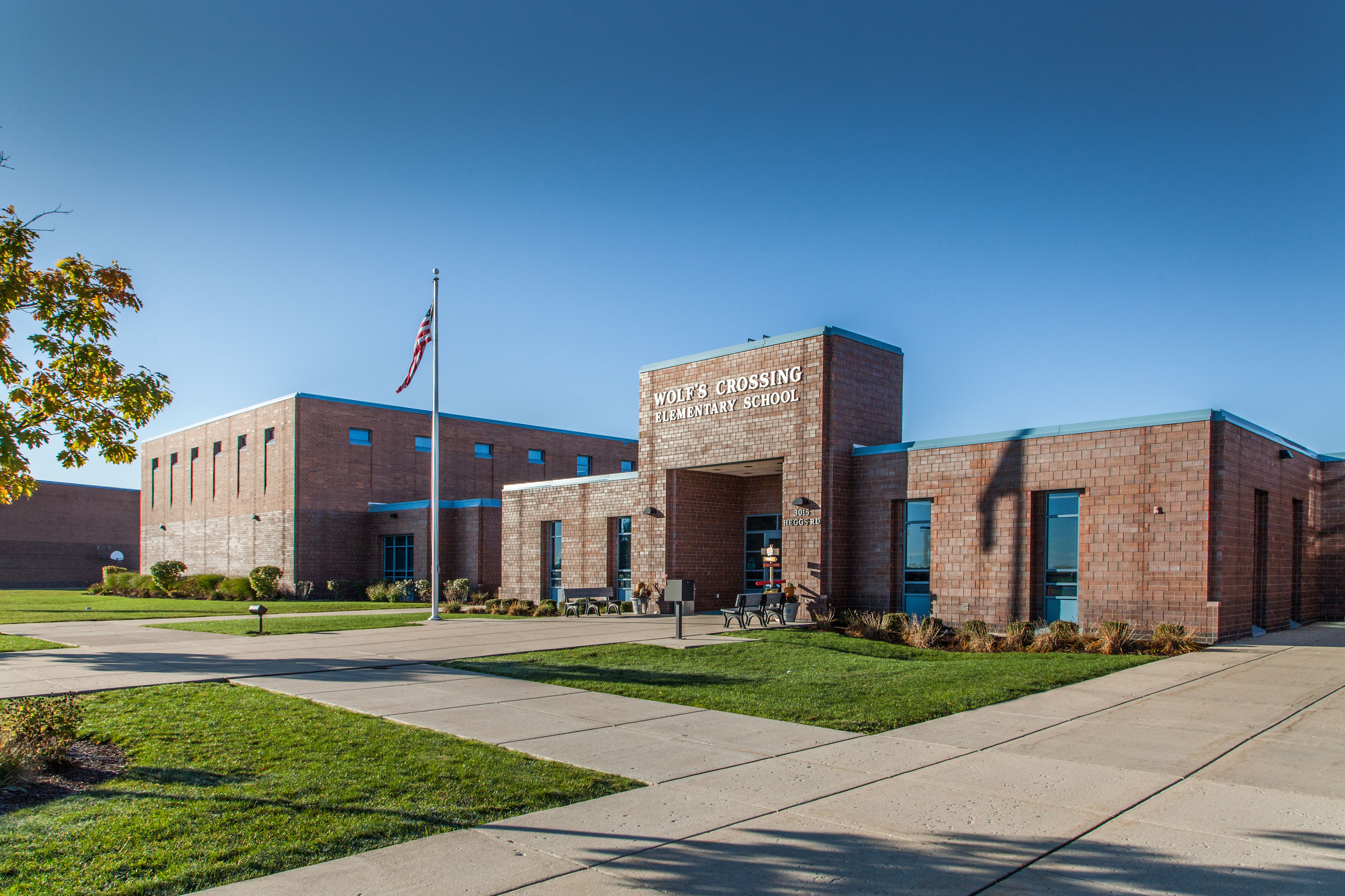 Wolf's Crossing building front showing multiple large sidewalks, benches in front of the main entrance, flagpole, and a blue sky behind the building. 